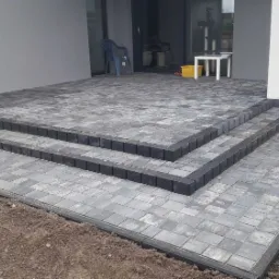 Ułożona kostka brukowa w odcieniach szarości tworząca schody wejściowe do domu z widocznymi fugami i obrzeżami z ciemnej kostki.