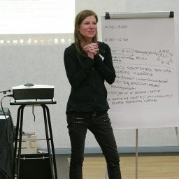 Grażyna Arendt GRAGA - Kobieta w ciemnym stroju stoi przed flipchartem z odręcznymi notatkami podczas prezentacji, z projektorem i laptopem w tle.