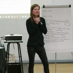 Kobieta w ciemnym stroju stoi przed flipchartem z odręcznymi notatkami podczas prezentacji, z projektorem i laptopem w tle.