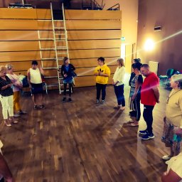 Grażyna Arendt GRAGA - Grupa osób ustawiona w okręgu na drewnianej podłodze, prawdopodobnie podczas szkolenia interpersonalnego, z drabiną opartą o ścianę w tle.