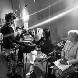 Grażyna Arendt GRAGA - Czarno-białe zdjęcie przedstawia młodą dziewczynę wręczającą plik dokumentów siedzącym dorosłym osobom podczas spotkania lub szkolenia w pomieszczeniu z drewnianą podłogą i oświetleniem ledowym...