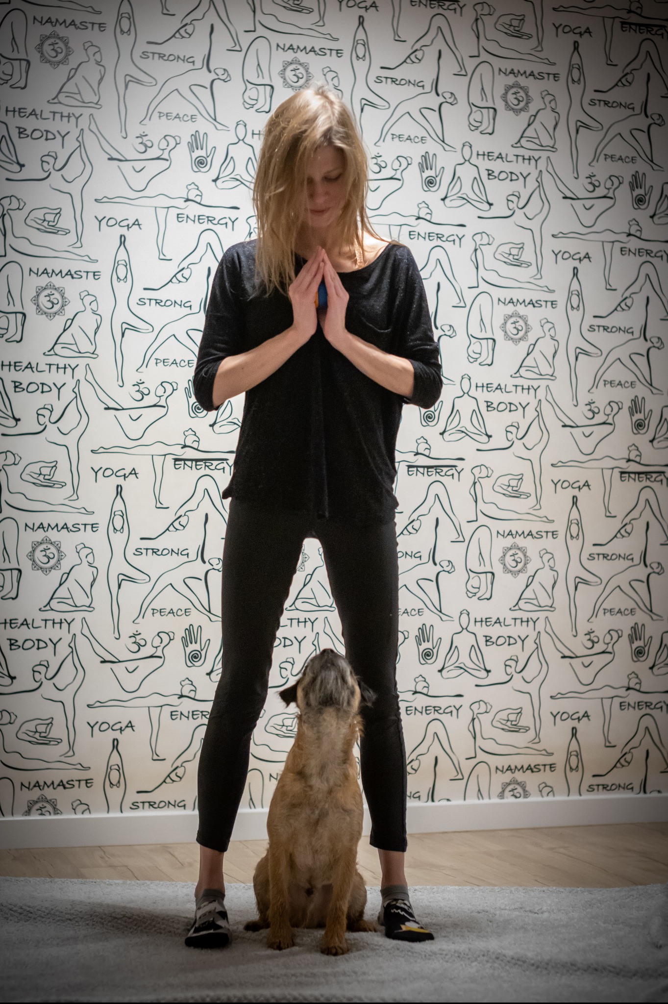 Kobieta w czarnym stroju do jogi z rękami złożonymi w geście modlitwy, stojąca na macie z psem siedzącym u jej stóp, na tle ściany z motywami jogi.
