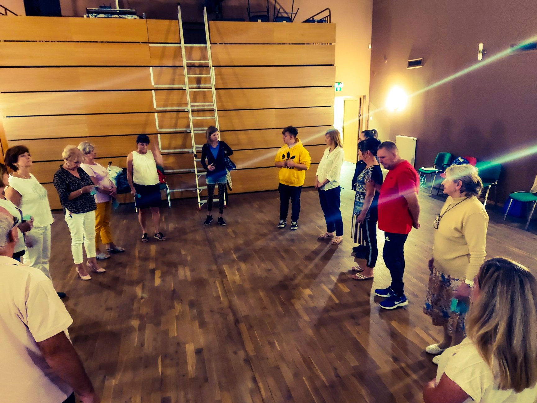 Grupa osób ustawiona w okręgu na drewnianej podłodze, prawdopodobnie podczas szkolenia interpersonalnego, z drabiną opartą o ścianę w tle.