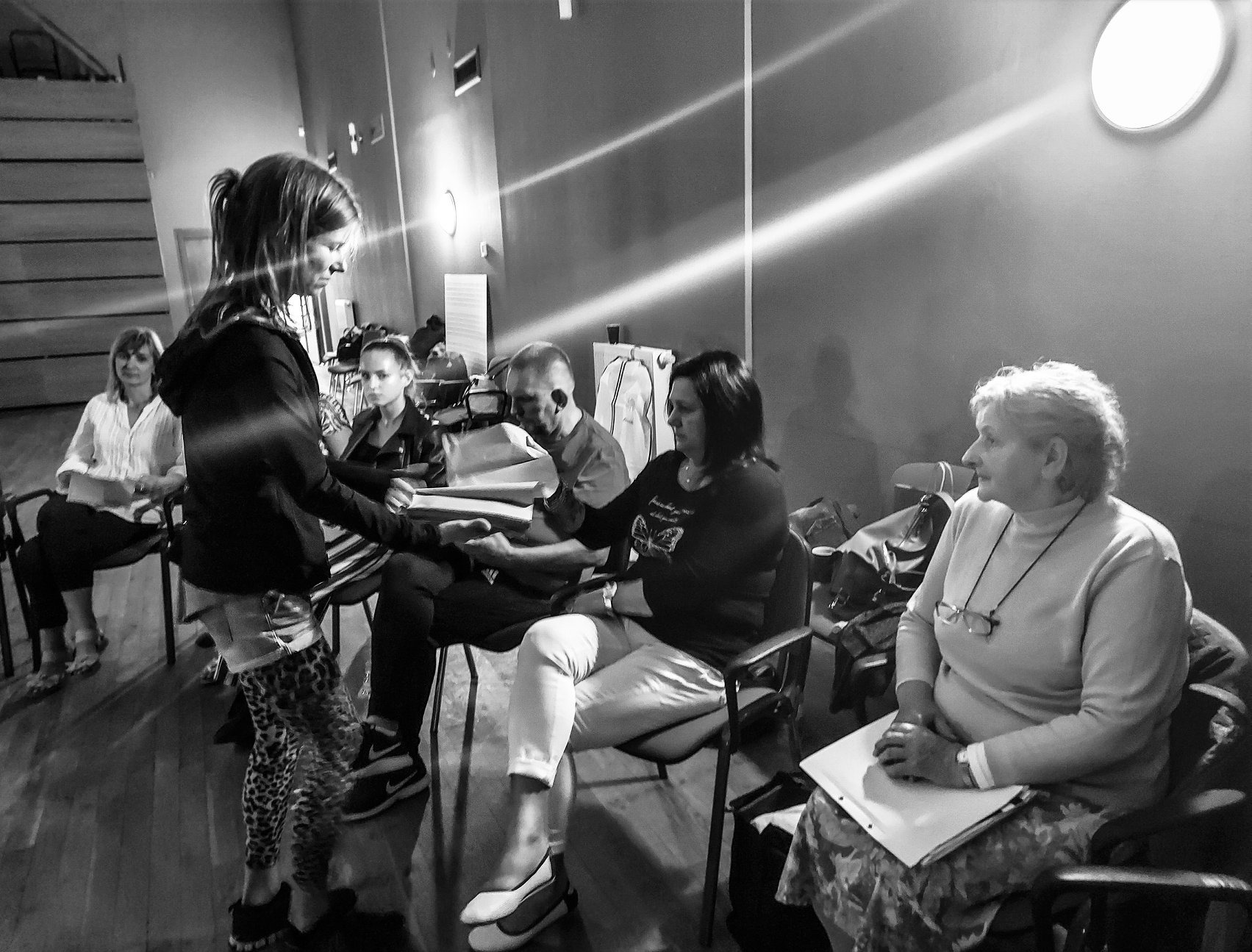 Czarno-białe zdjęcie przedstawia młodą dziewczynę wręczającą plik dokumentów siedzącym dorosłym osobom podczas spotkania lub szkolenia w pomieszczeniu z drewnianą podłogą i oświetleniem ledowym...