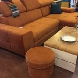 Pomarańczowy narożnik z regulowanymi zagłówkami, obok pufa i stolik kawowy z dekoracyjnym obrusem. Na narożniku wzornik tkanin. W tle elementy wyposażenia sklepu.