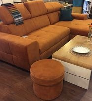 Pomarańczowy narożnik z regulowanymi zagłówkami, obok pufa i stolik kawowy z dekoracyjnym obrusem. Na narożniku wzornik tkanin. W tle elementy wyposażenia sklepu.