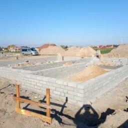 Fundamenty domu z bloczków betonowych, widoczne zarysy ścian, przygotowane pod dalszą budowę, w tle hałdy piasku i zabudowa jednorodzinna.
