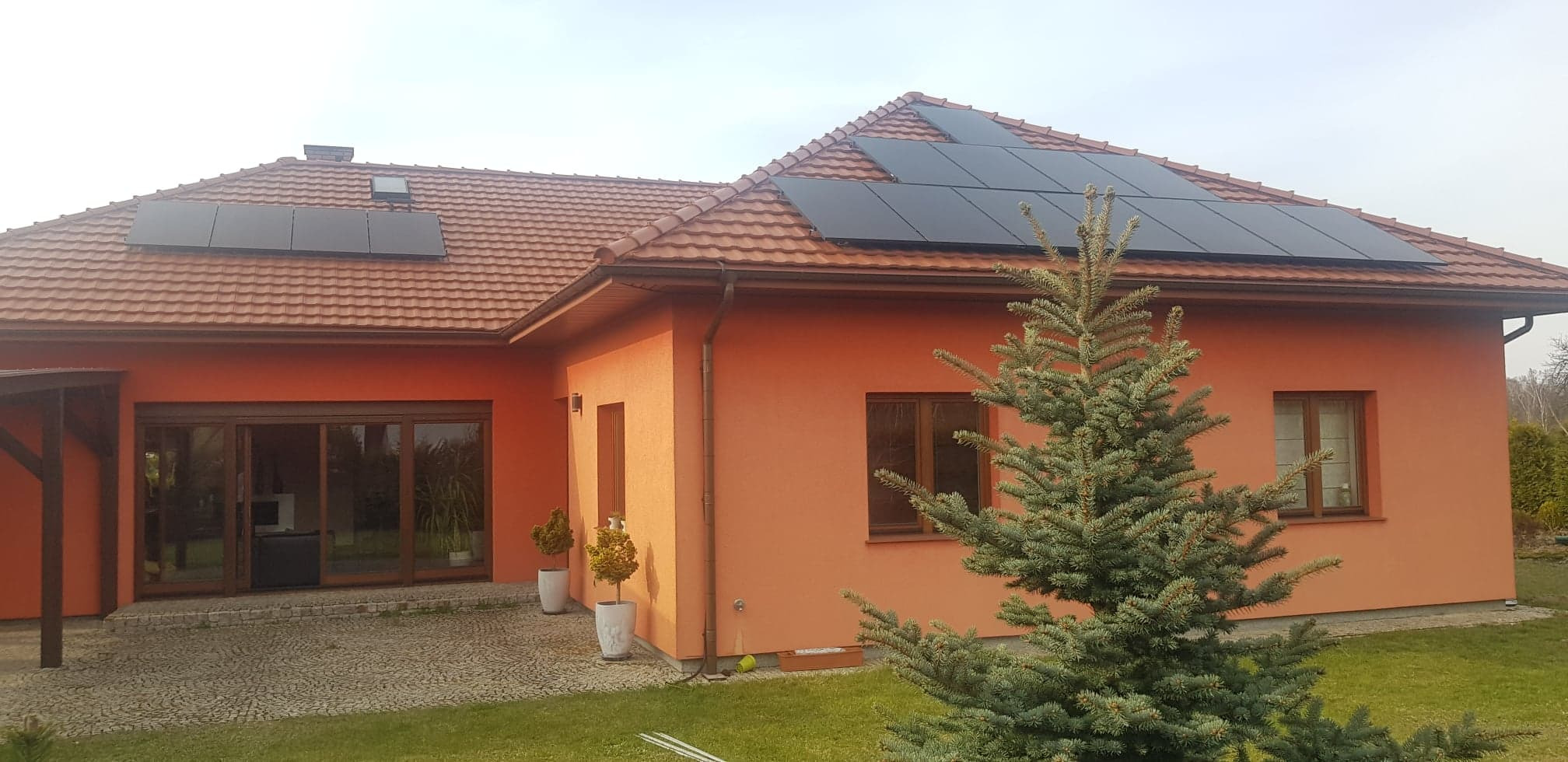 Pomarańczowy dom jednorodzinny z brązową dachówką i panelami słonecznymi na dachu, widok z poziomu trawnika, z choinką na pierwszym planie.
