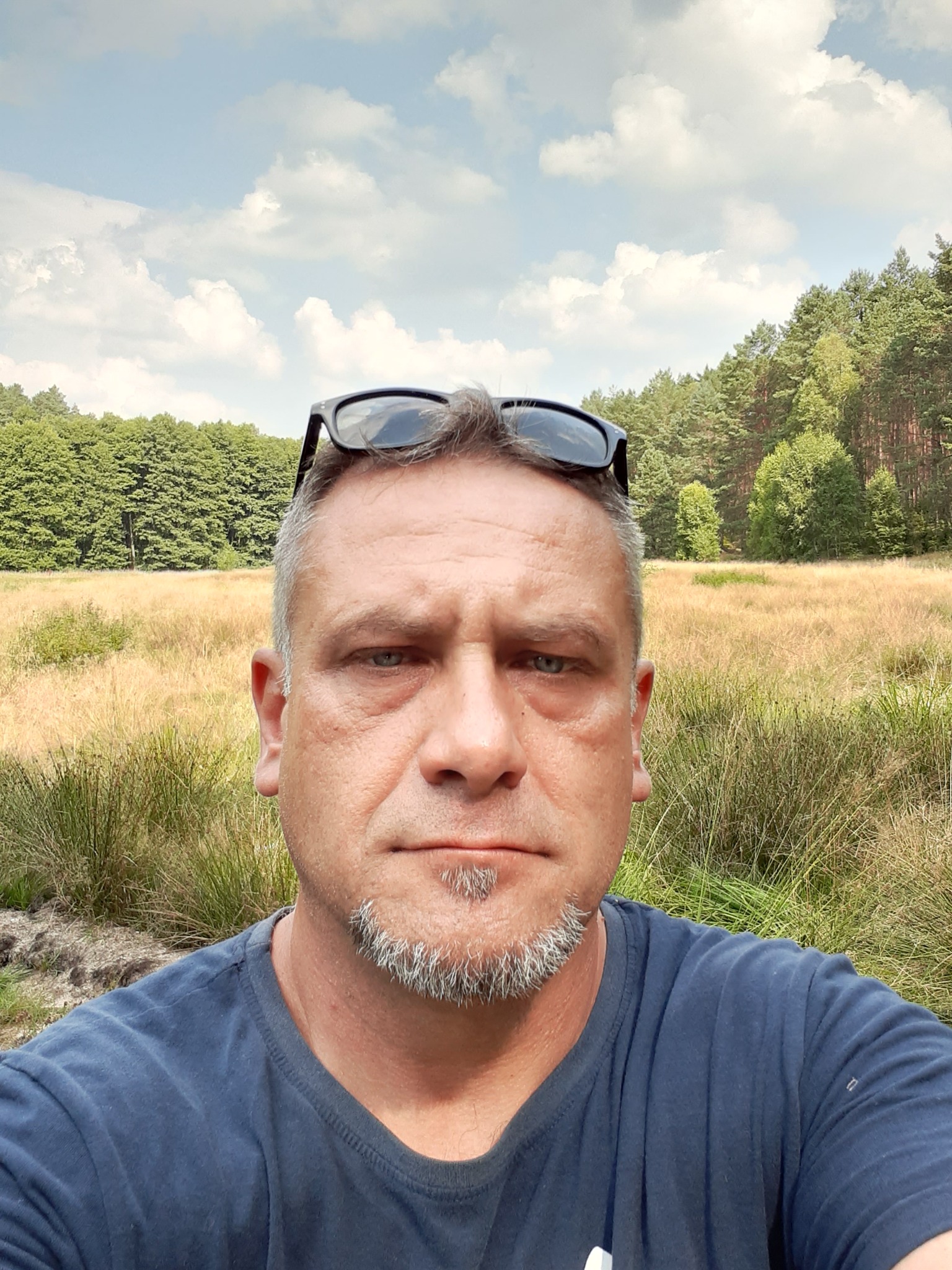 Autoportret mężczyzny z okularami na głowie, na tle łąki i lasu w słoneczny dzień.