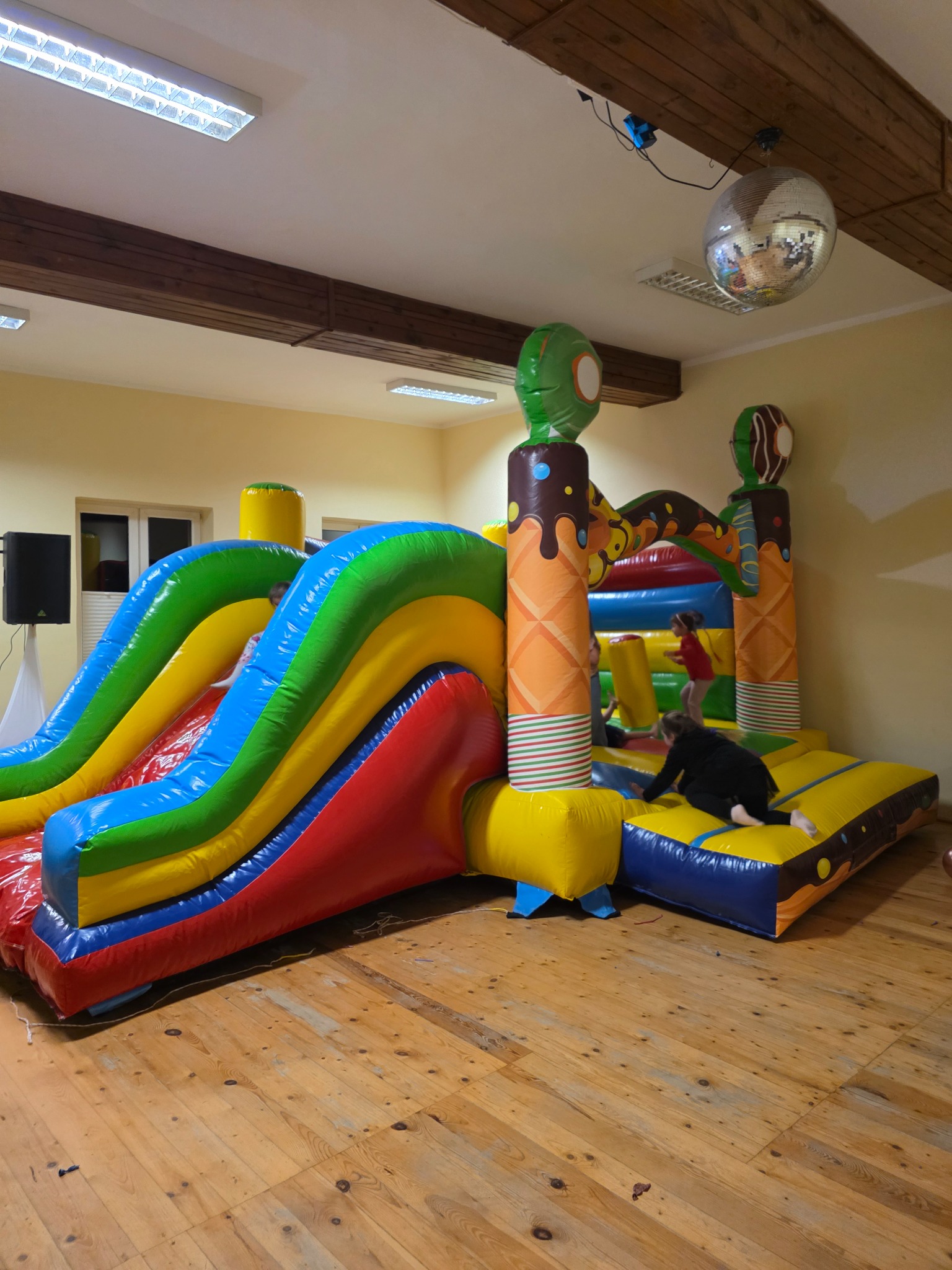 Kolorowy dmuchany zamek w kształcie lodów i zjeżdżalni, dzieci bawią się w środku. W tle parkiet, lampy i kula dyskotekowa.