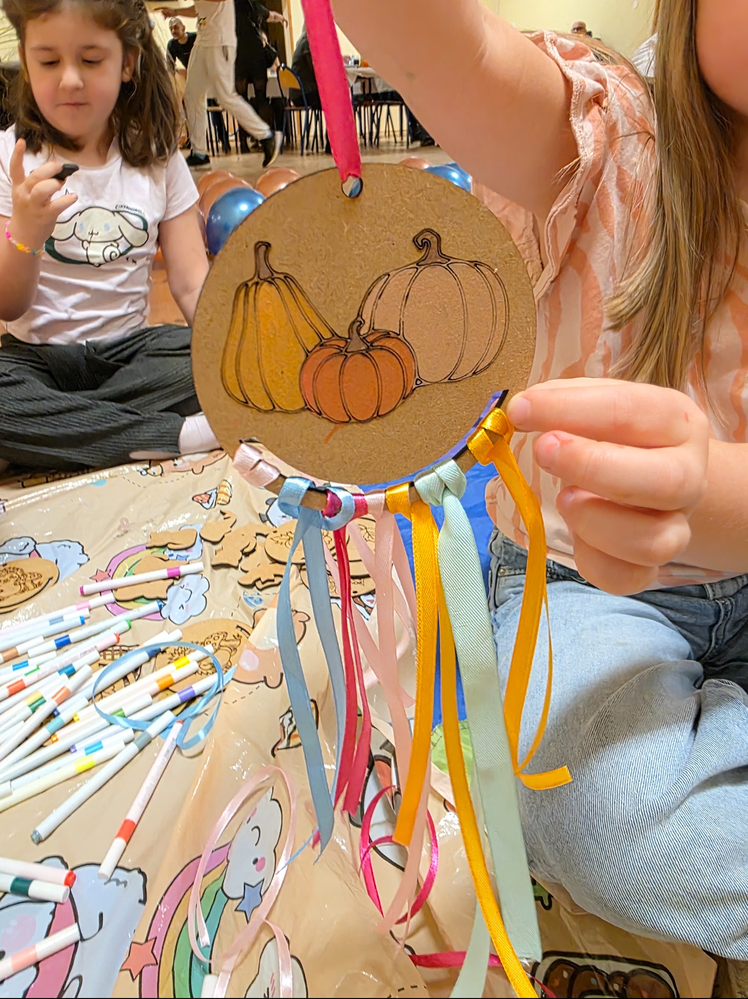 Dziecko tworzy jesienną dekorację z dyniami i kolorowymi wstążkami na warsztatach artystycznych, siedząc na podłodze z mazakami i innymi dziećmi w tle.
