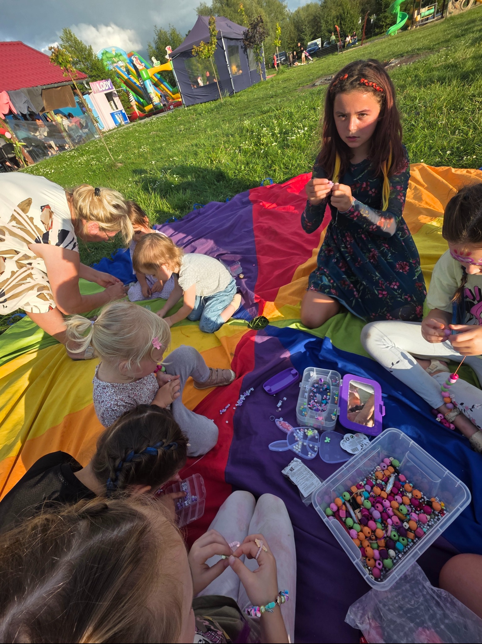 Dzieci na kolorowej chuście tworzą biżuterię z koralików. W tle dmuchany zamek i namiot, sugerujące imprezę plenerową. Dziewczynka z dredami patrzy w obiektyw.