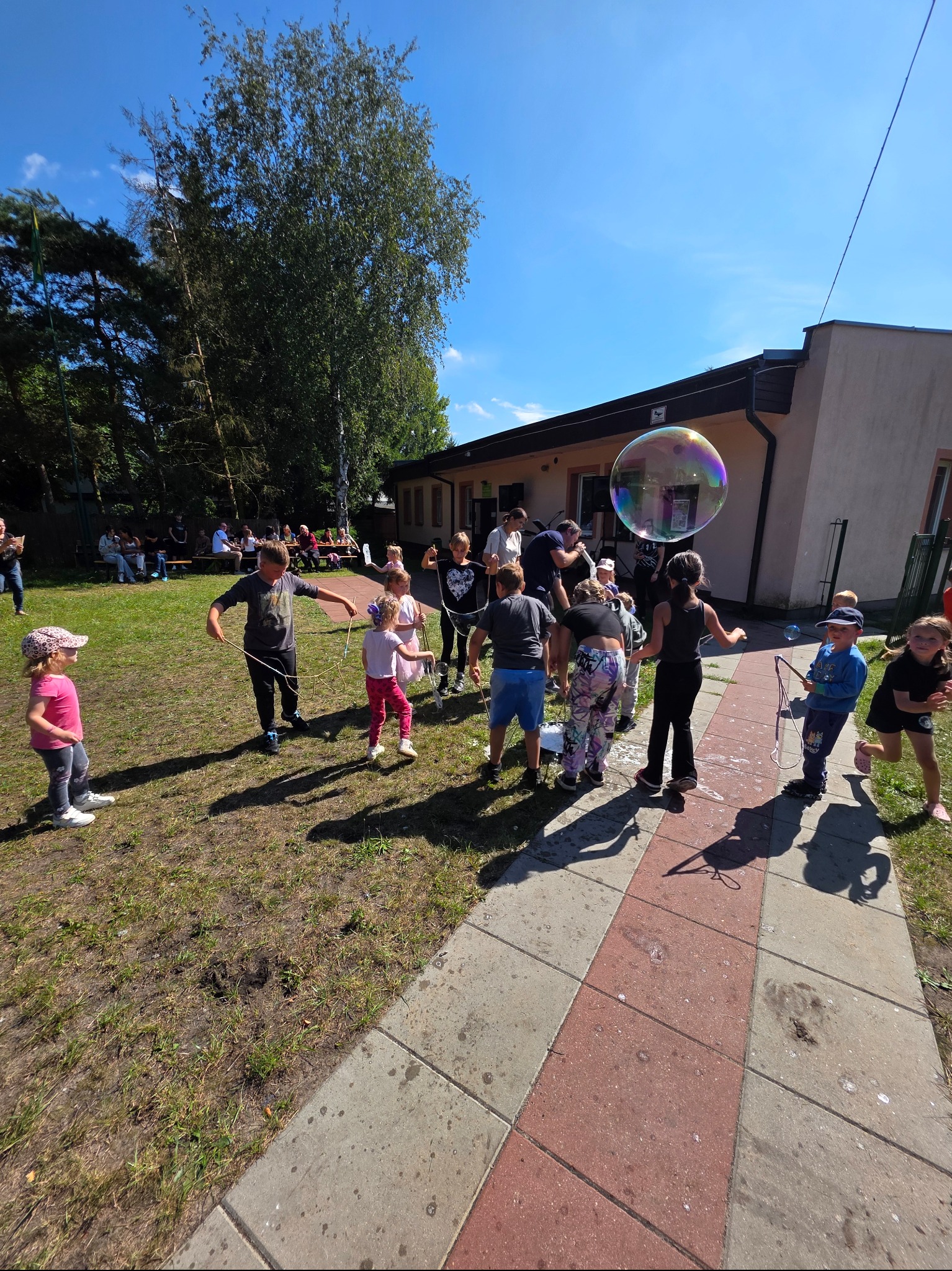 Dzieci bawią się na trawie, tworząc ogromne bańki mydlane. W tle budynek i drzewa, słoneczny dzień. Zabawa na świeżym powietrzu.