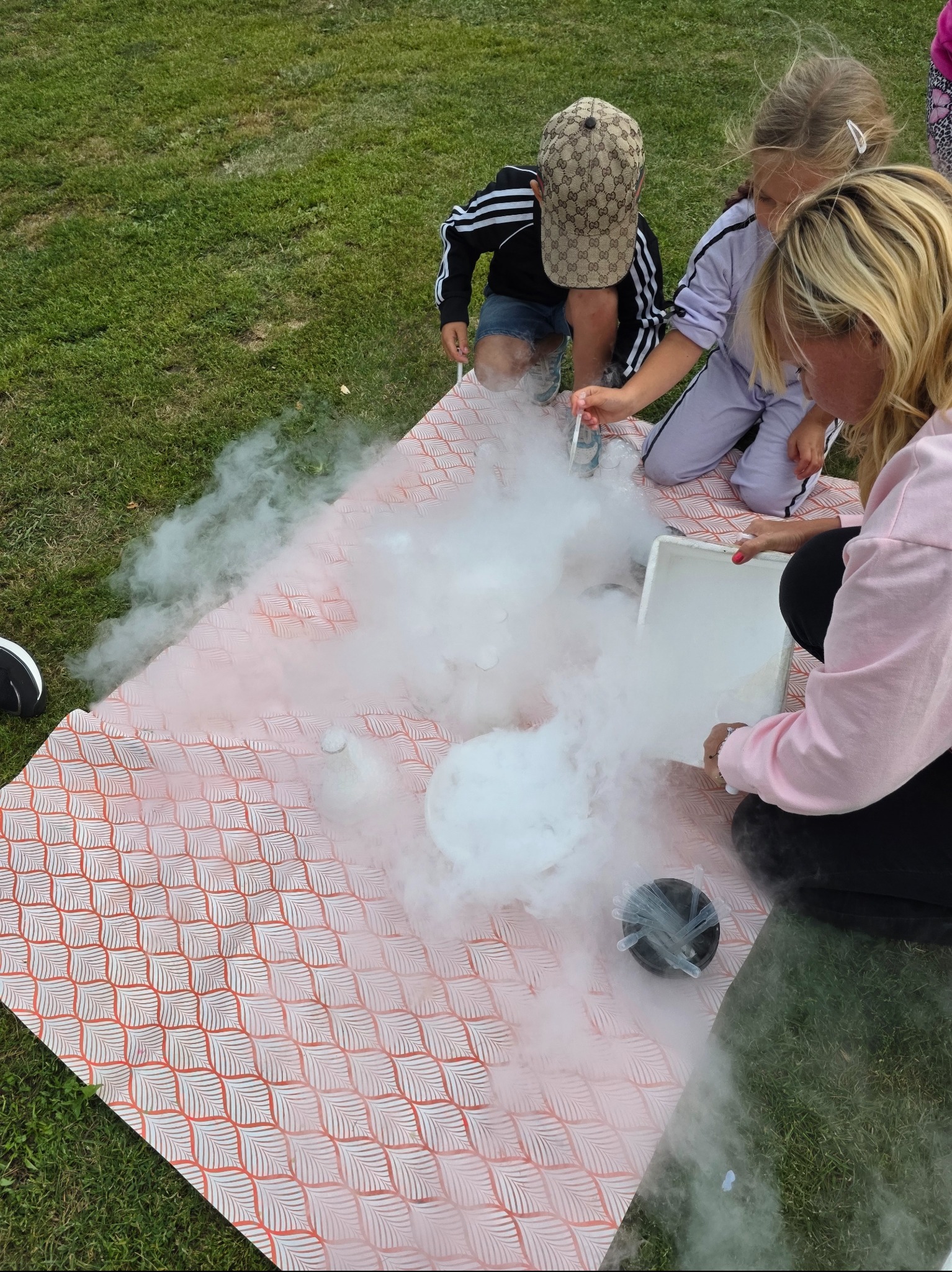Pokaz naukowy z suchym lodem na trawie: dzieci obserwują eksperyment z dymiącą substancją, prowadzony przez osobę w różowej bluzie. Piknikowa atmosfera.