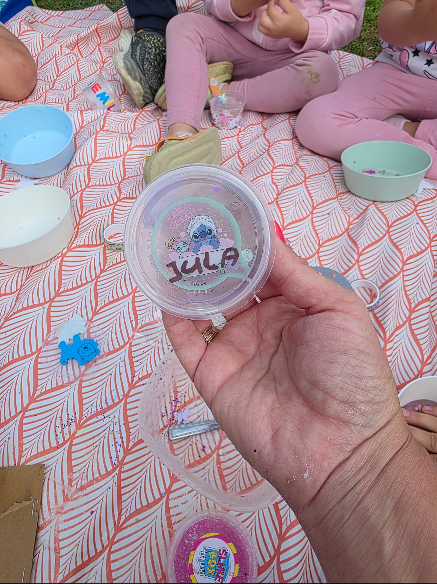 Ręka trzymająca pojemnik ze slime z imieniem 'Jula' na pikniku z dziećmi, miseczki i brokat na kocu w liście. Kreatywne warsztaty dla dzieci.