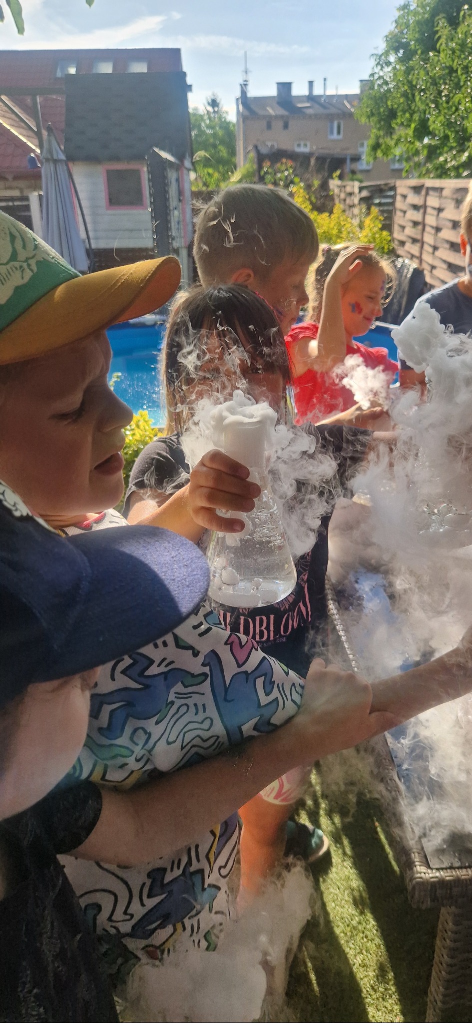 Dzieci obserwują eksperyment z dymem podczas animacji na świeżym powietrzu. W tle basen i drewniany płot. Uwagę przykuwa reakcja dzieci na efektowny pokaz.