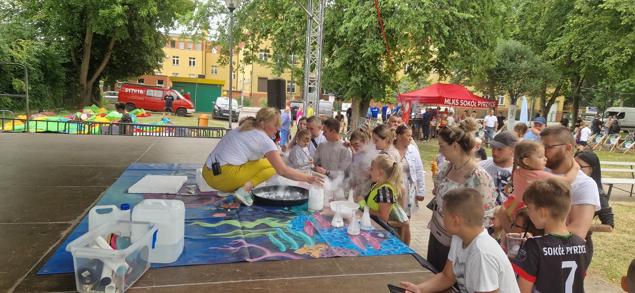 Animator w żółtych spodniach prezentuje eksperymenty z dymem dla dzieci na scenie plenerowej. W tle dmuchany zamek i budynki.