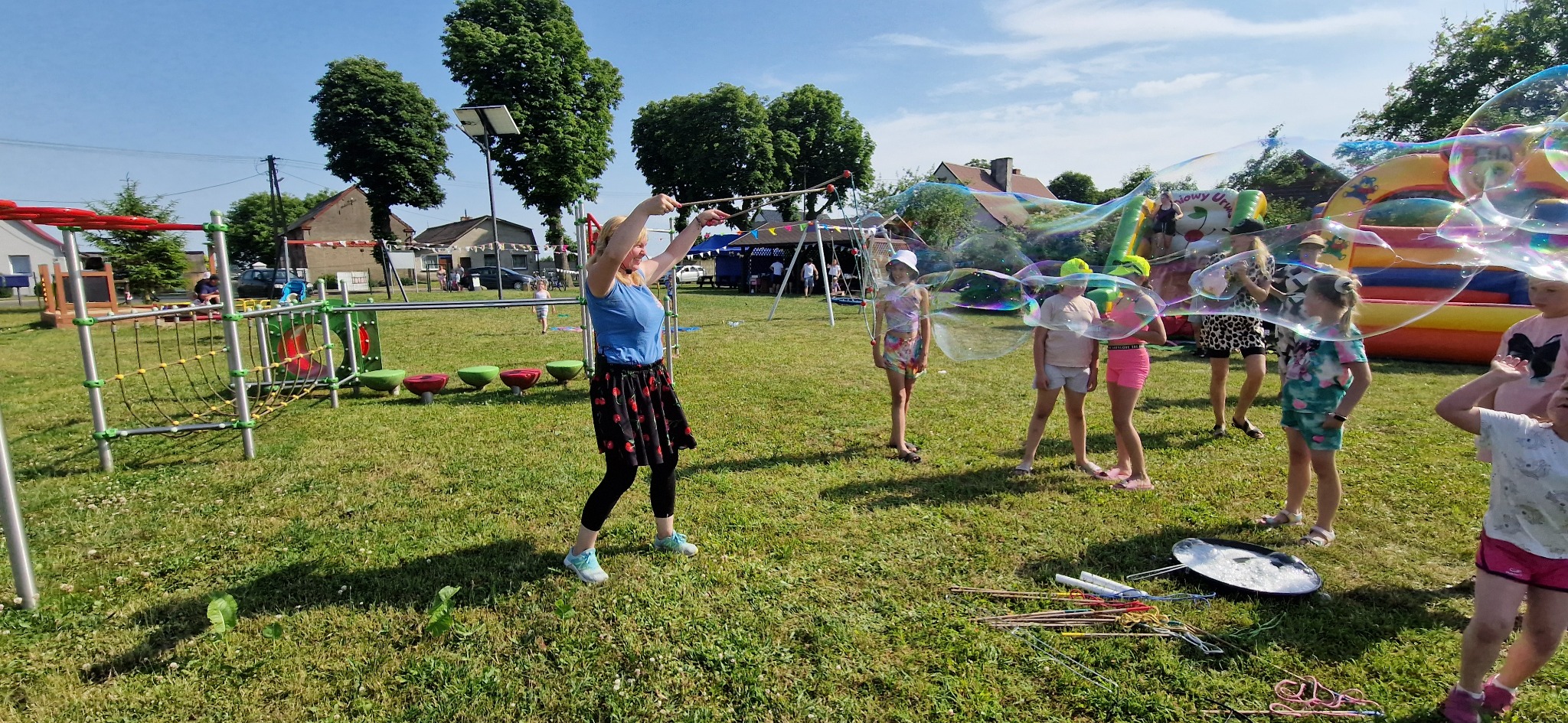 Animator puszcza gigantyczne bańki mydlane na trawie, otoczony grupą dzieci podczas imprezy plenerowej. W tle dmuchany zamek i plac zabaw.