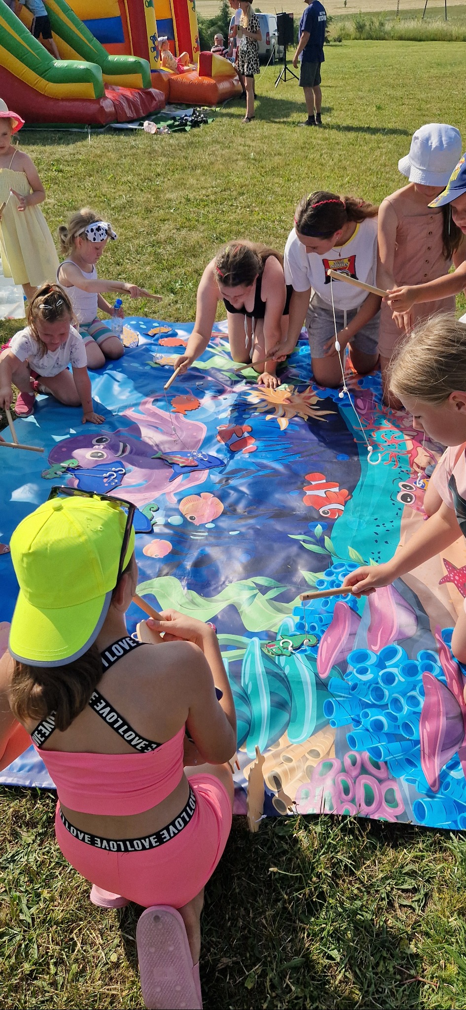 Dzieci bawią się w łowienie rybek na kolorowej macie z motywem morskim. W tle dmuchany zamek i osoby dorosłe. Zabawa na świeżym powietrzu.