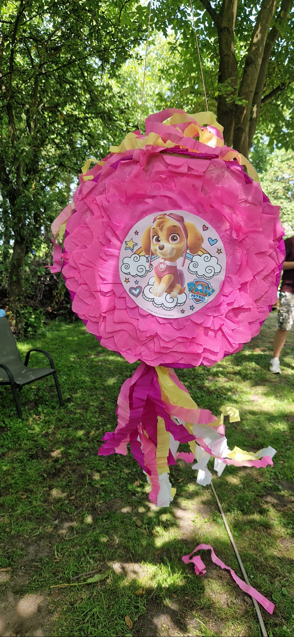 Ręcznie robiona, różowa piniata z wizerunkiem Skye z Psiego Patrolu, zawieszona na zewnątrz. Dekoracja urodzinowa dla dziecka, widok z dołu, trawa i drzewa w tle.