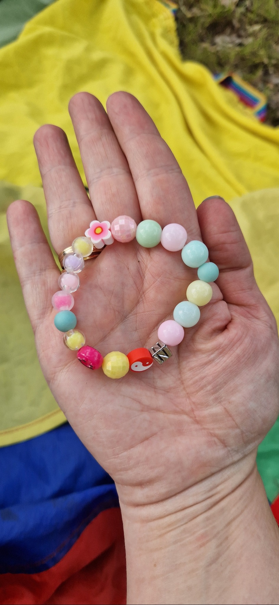 Dłoń prezentuje kolorową bransoletkę z koralików i motywem kwiatka na tle żółtej tkaniny. Bransoletka w pastelowych odcieniach, idealna na dziecięce przyjęcie.