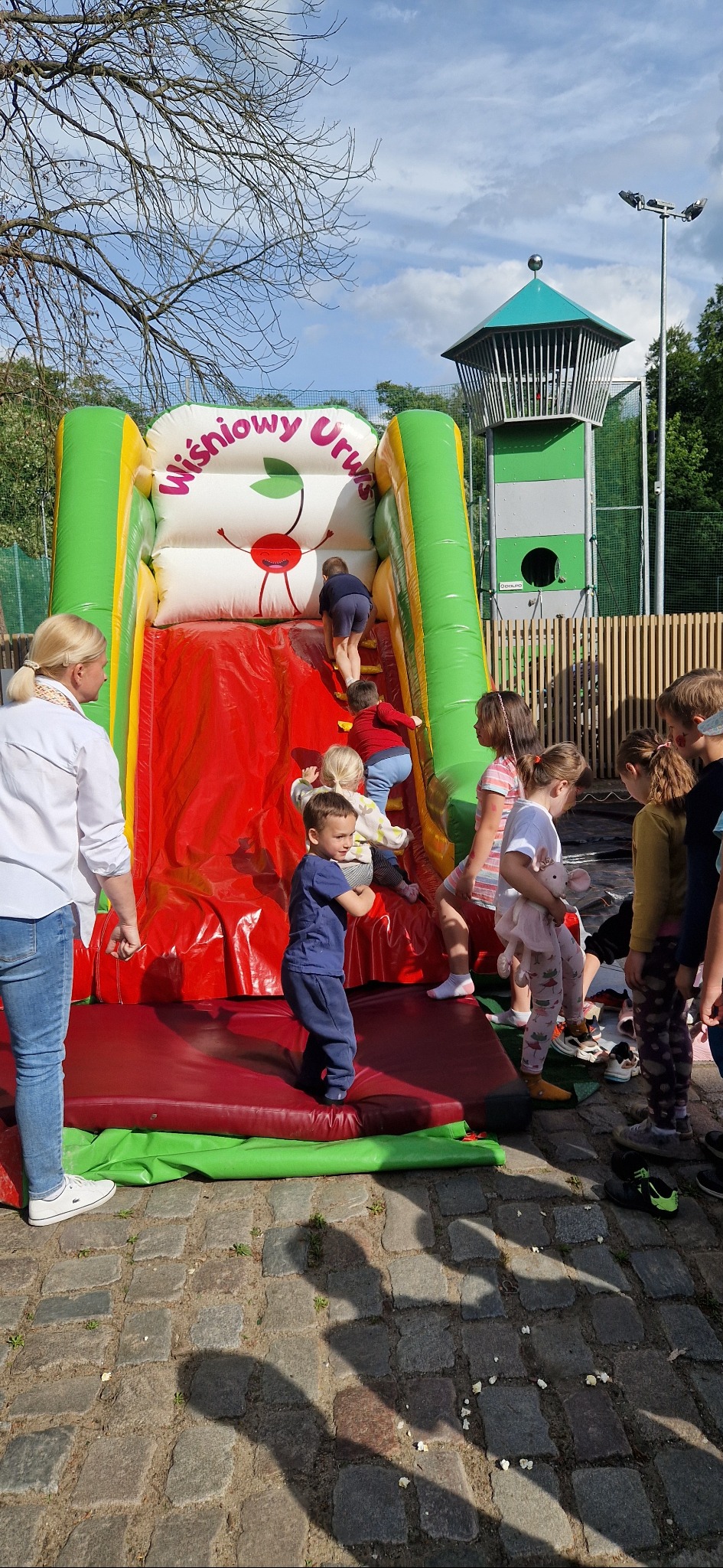 Dzieci wspinają się i zjeżdżają z dmuchanej zjeżdżalni w kształcie wiśni na tle zieleni i wieży obserwacyjnej. Widoczne cienie na brukowanej nawierzchni.