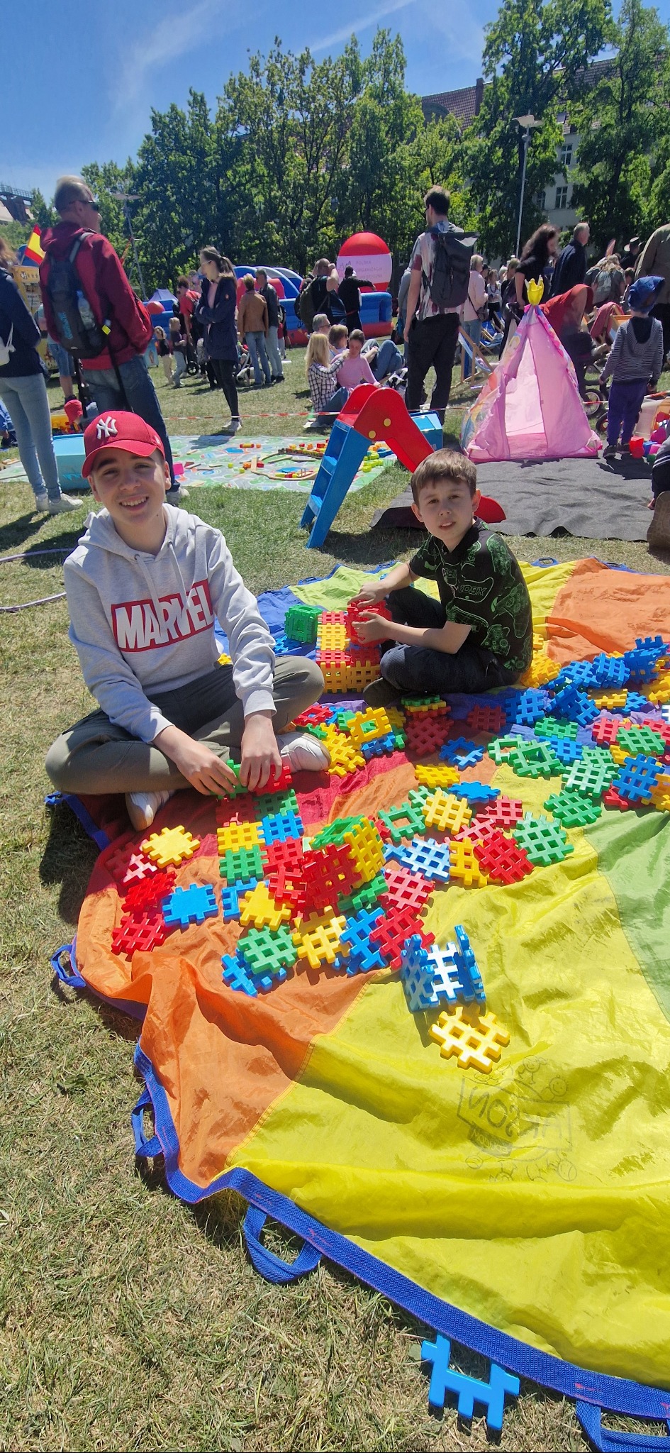 Dwóch chłopców siedzi na trawie, bawiąc się kolorowymi klockami na tle pikniku z dmuchańcami i innymi dziećmi w słoneczny dzień.