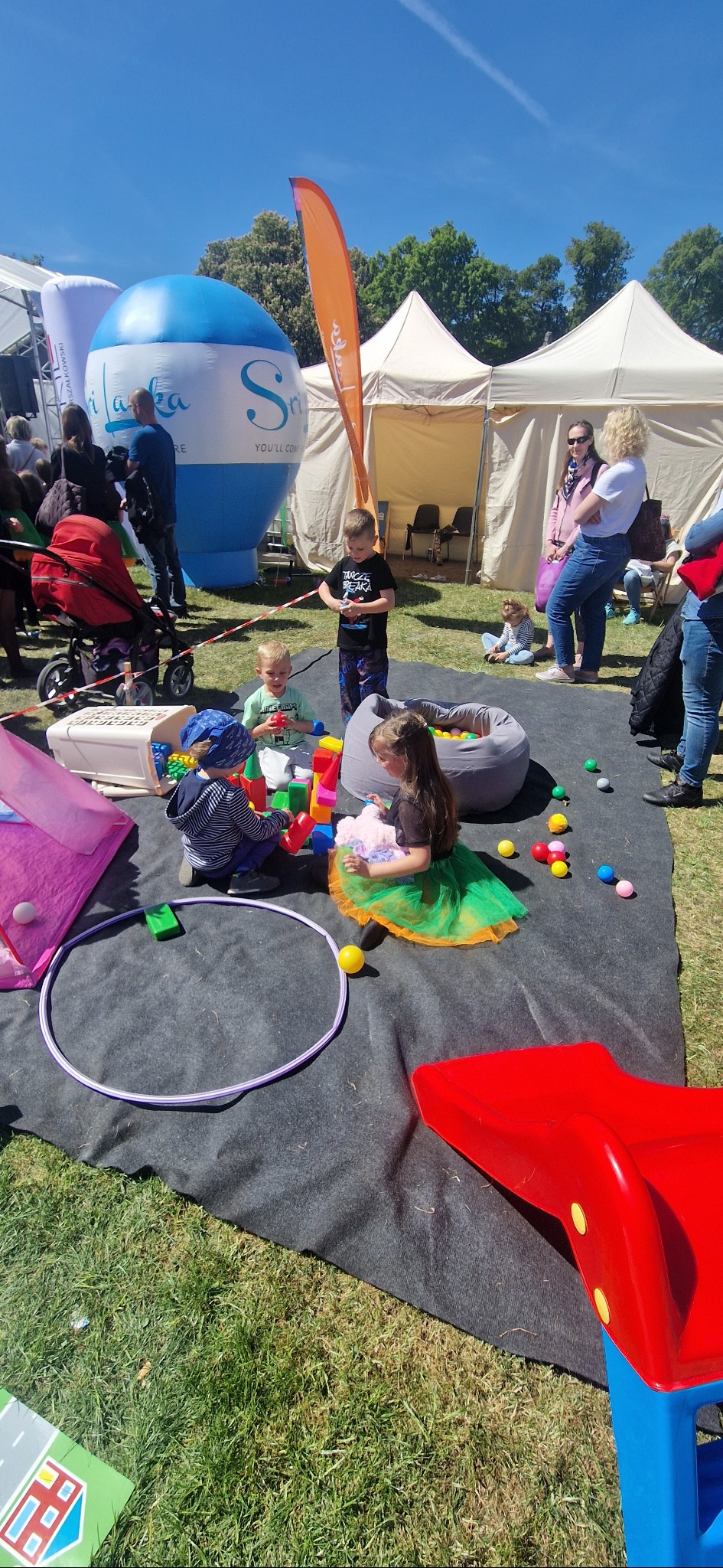 Dzieci bawią się klockami i piłkami na czarnej macie podczas plenerowej imprezy z dmuchanym balonem i namiotami w tle, w słoneczny dzień.