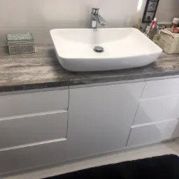 Nowoczesna łazienka z białą, kwadratową umywalką nablatową na blacie imitującym drewno i białymi szafkami z szufladami, uzupełniona o dekoracyjne akcesoria.