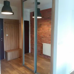 Przedpokój z dużą szafą przesuwną z lustrami, drewnianym panelem z wieszakami i siedziskiem, oświetlony wiszącą lampą, z podłogą imitującą drewno i ścianą z czerwonej cegły widoczną w lustrze.