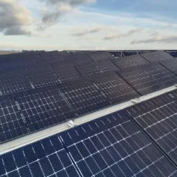 Rozległa instalacja paneli słonecznych na dachu, widoczna z góry, z tłem błękitnego nieba z chmurami i odległym krajobrazem.