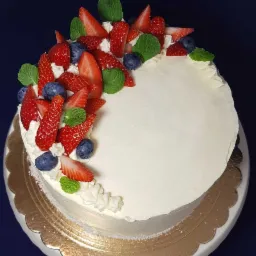 Tort z białym kremem, udekorowany świeżymi truskawkami, borówkami i listkami mięty, umieszczony na złotej podkładce.