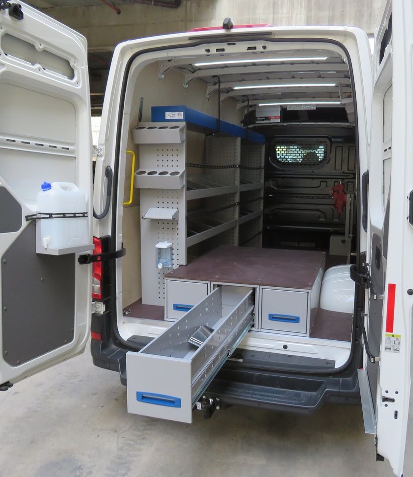 Wnętrze białego furgonu z otwartymi tylnymi drzwiami, wyposażone w system regałów i szuflad, wysuwana szuflada na zewnątrz, kanister z wodą przy drzwiach, oświetlenie LED wewnątrz.