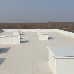 Biała, płaska powierzchnia dachu pokryta membraną z widocznymi świetlikami, w tle las i zabudowa jednorodzinna.