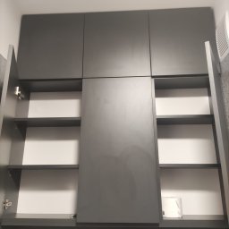 MebleBieniek.pl - Szara szafka wisząca z otwartymi drzwiczkami ukazującymi puste półki, na dolnej półce widoczny zwój papieru toaletowego, na dole dwie białe ceramiczne figurki ptaków.