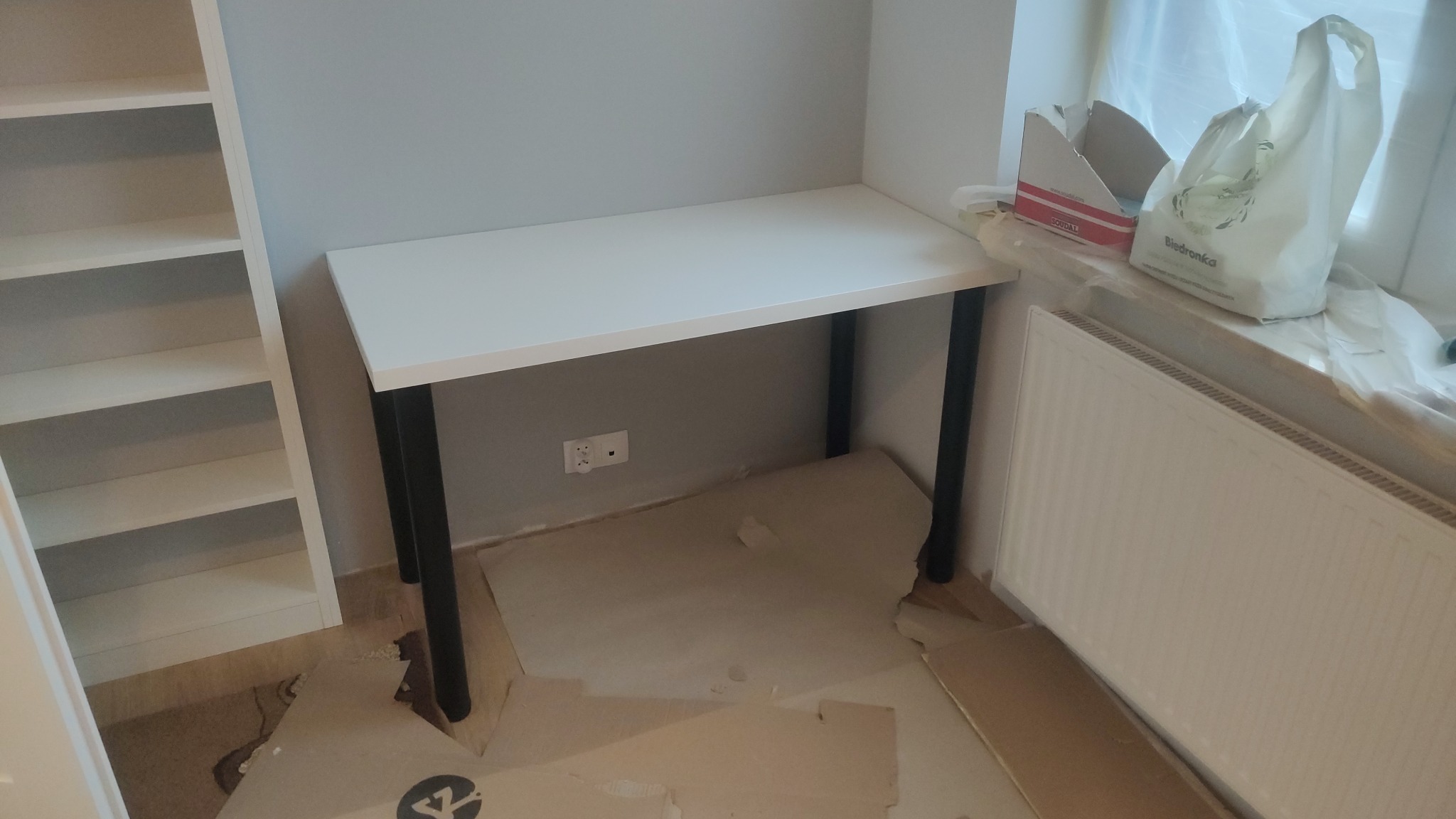 Białe biurko z czarnymi nogami ustawione przy ścianie obok regału, zabezpieczone kartonem podłoga w trakcie remontu, widoczny grzejnik i okno.