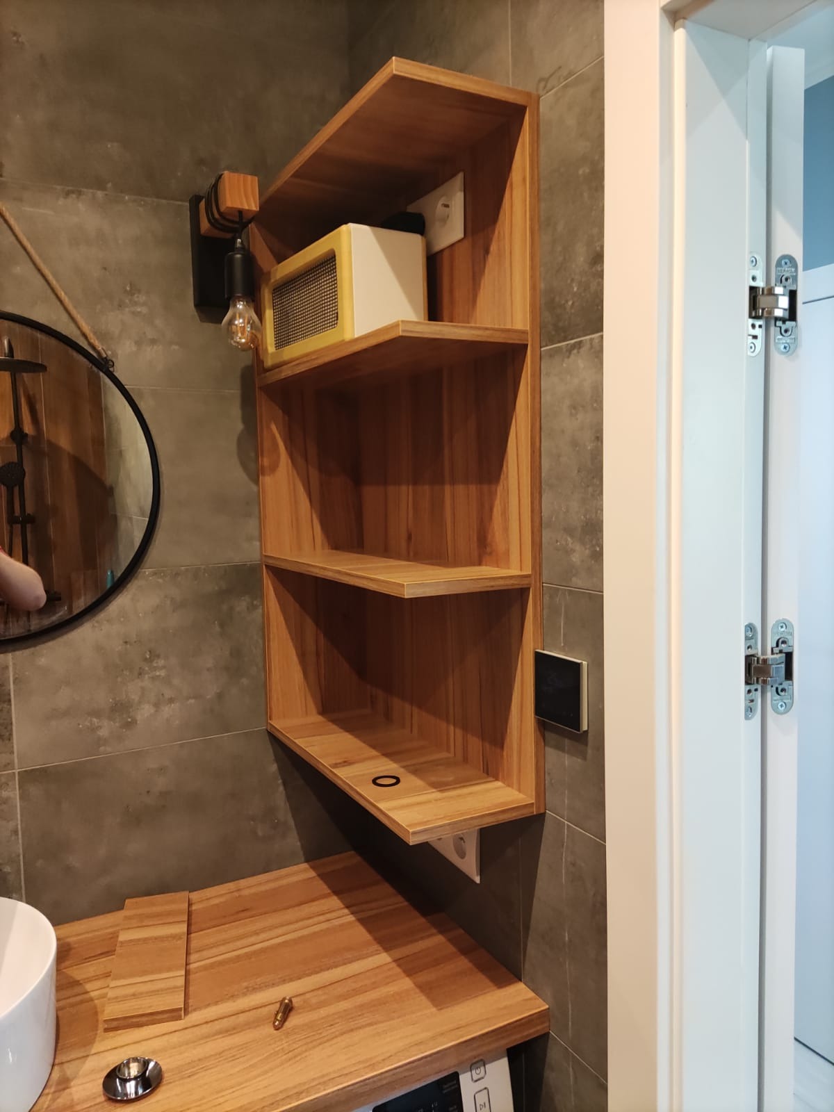 Drewniana półka narożna w łazience, z zaokrąglonym blatem, obok drzwi z ukrytymi zawiasami, oświetlona żarówką edisona i uzupełniona radiem w stylu retro.