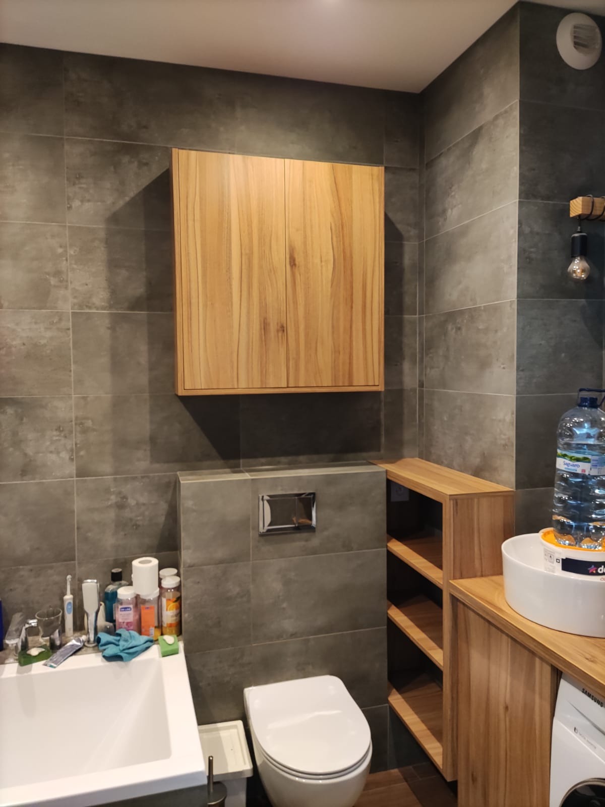 Wnętrze łazienki z drewnianą szafką wiszącą nad toaletą, drewnianym regałem i umywalką nablatową, szare płytki na ścianach, oświetlenie dekoracyjne.