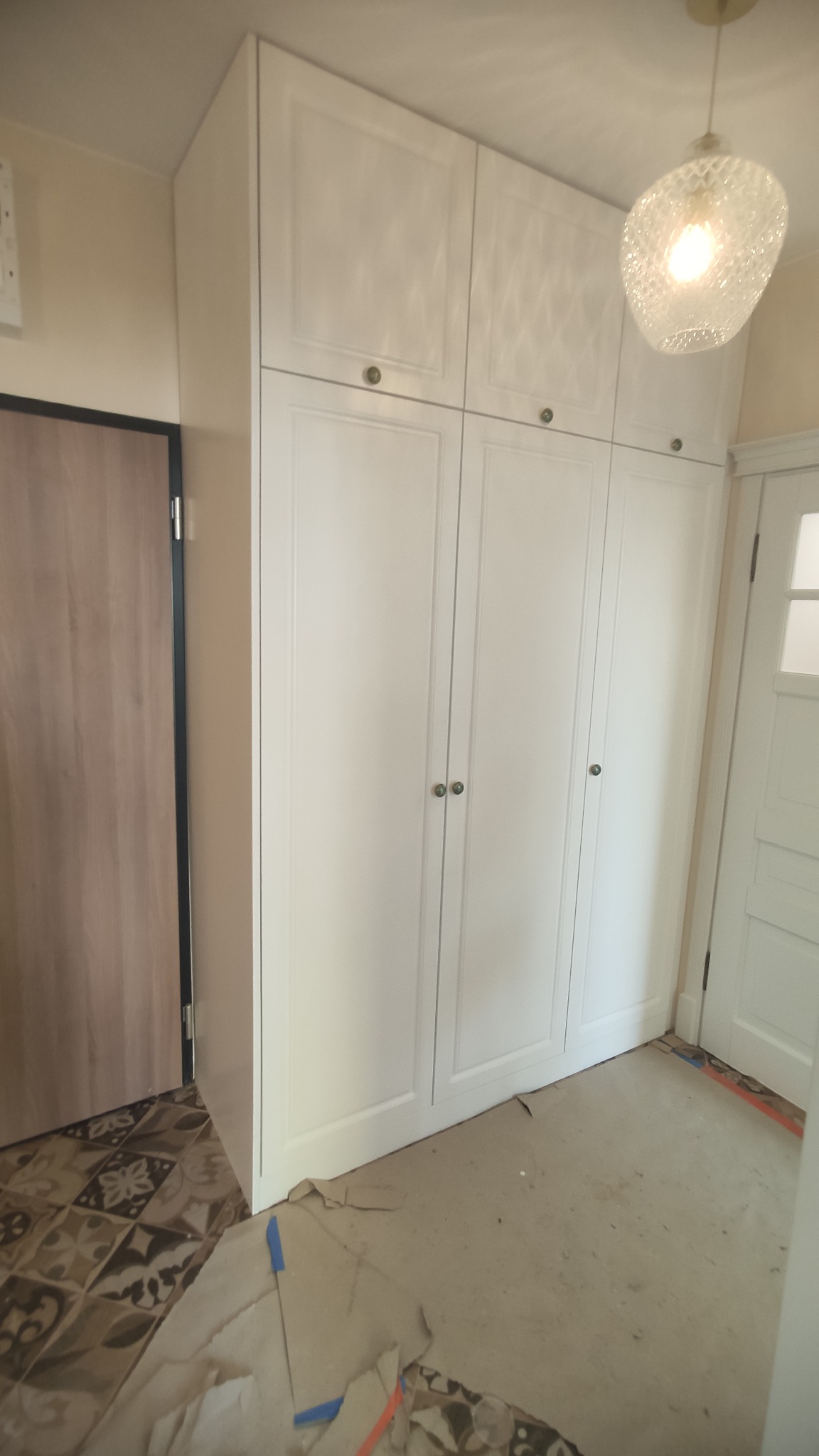 Wysoka, biała zabudowa szafowa z czterema parami drzwi z okrągłymi uchwytami, widoczna w trakcie prac wykończeniowych, z fragmentami zabezpieczeń podłogi i sąsiednich drzwi.