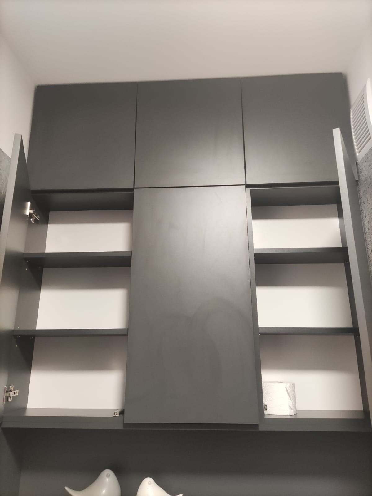 Szara szafka wisząca z otwartymi drzwiczkami ukazującymi puste półki, na dolnej półce widoczny zwój papieru toaletowego, na dole dwie białe ceramiczne figurki ptaków.