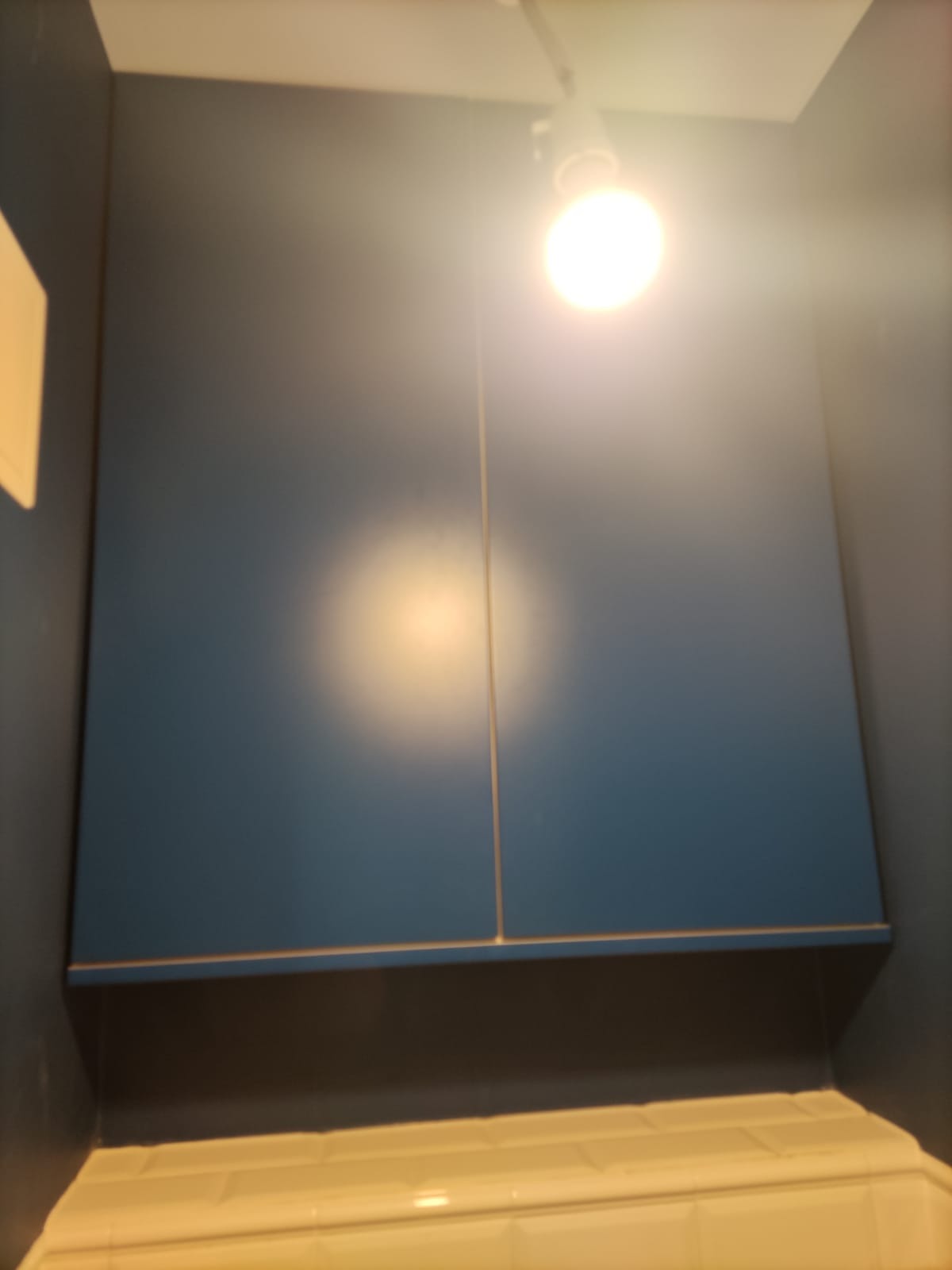 Niebieska szafka wisząca z dwoma drzwiczkami, oświetlona od góry punktowym światłem, zamontowana nad białymi płytkami w kuchni.