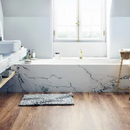 Jasna łazienka z marmurową obudową wanny, drewnianą podłogą i złotą armaturą, widok z niskiego kąta na szary dywanik.
