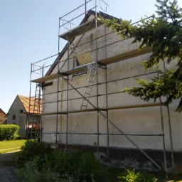 Dom jednorodzinny w trakcie remontu elewacji, otoczony rusztowaniem i zielenią.