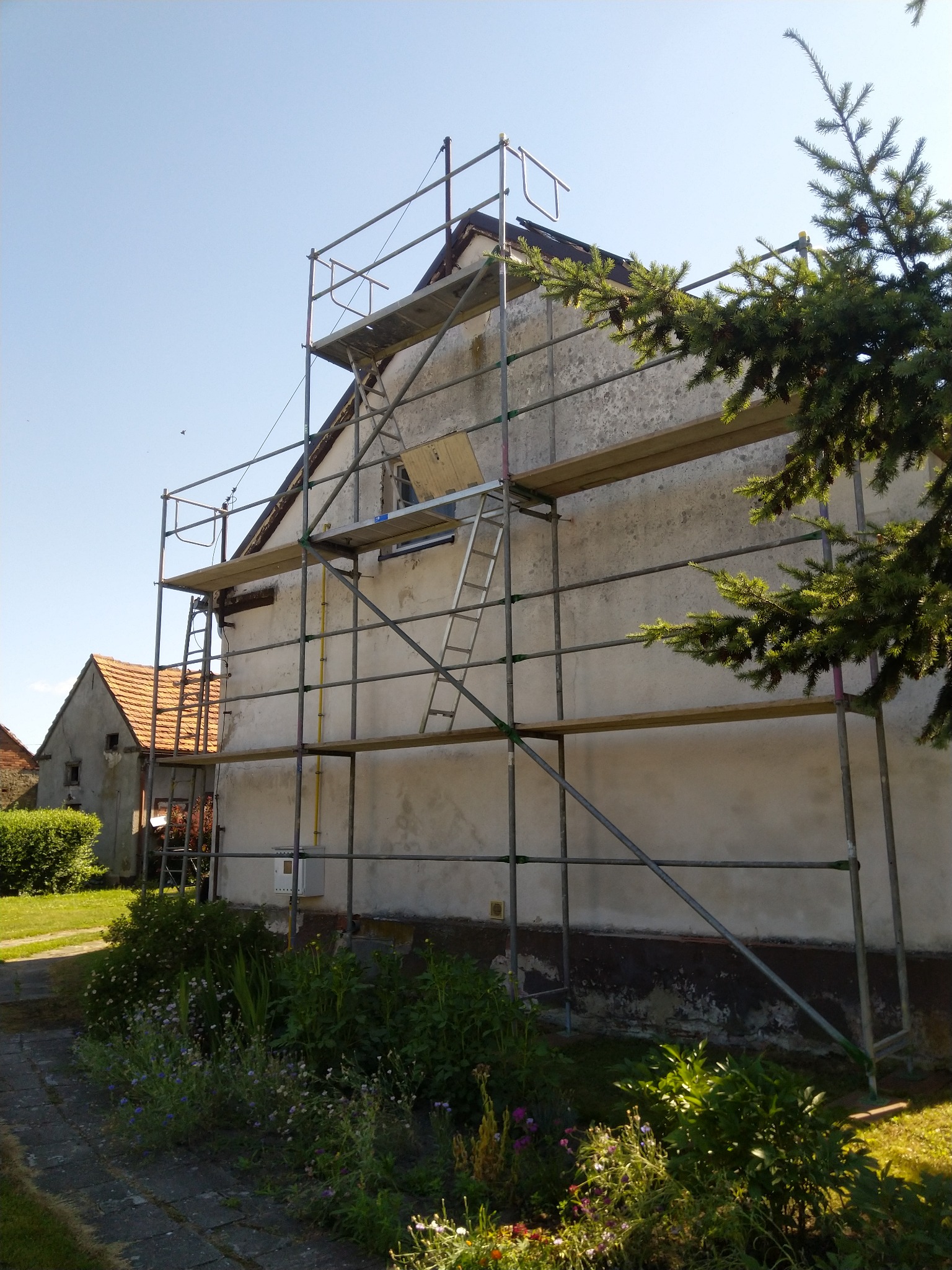 Dom jednorodzinny w trakcie remontu elewacji, otoczony rusztowaniem i zielenią.