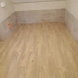 Wykończona łazienka z drewnopodobną podłogą, jasnymi płytkami ściennymi i instalacją hydrauliczną w rogu.