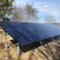 Zestaw paneli słonecznych na metalowej konstrukcji, zamontowany na trawiastym terenie z drzewami w tle, widoczne mocowania paneli.