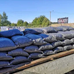 Rząd work&oacute;w z opałem, czarne worki ułożone na pace samochodu dostawczego, w tle tablica informacyjna składu opału i błękitne niebo.