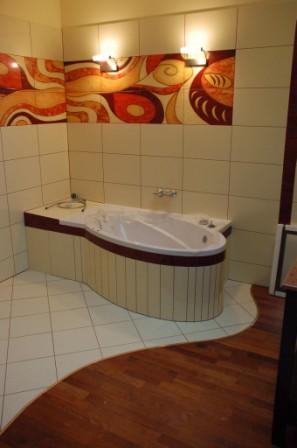 Wanna z hydromasażem w łazience wykończonej płytkami z wzorem i drewnianą podłogą, oświetlona przez dwa kinkiety.