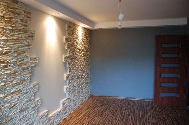 Pokój po remoncie z dekoracyjną ścianą wyłożoną kamieniem i parkietem ułożonym w jodełkę, widoczna drewniana stolarka drzwiowa z przeszkleniami.