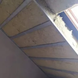 Skośne poddasze w trakcie izolacji, widoczne metalowe profile, wełna mineralna i fragment okna dachowego. Na podłodze leżą rolki wełny izolacyjnej w czerwonych opakowaniach.